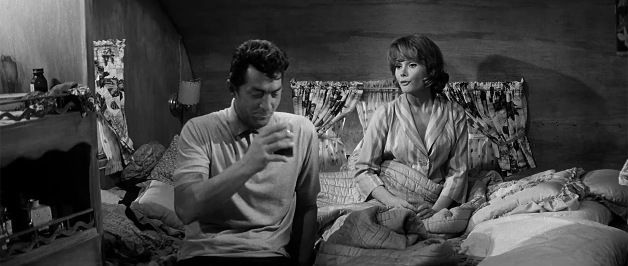 Kiss Me, Stupid (1964) movie still: medium — A cramped, dimly lit trailer interior features wood paneled walls and floral patterned curtains. A man…