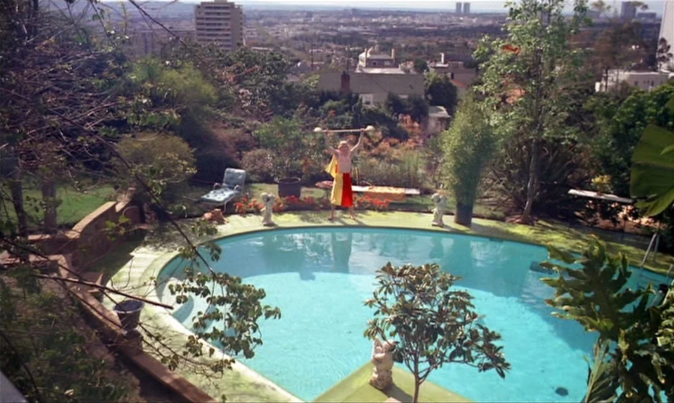 Lions Love (1969) movie still: wide — A sunny hillside swimming pool overlooks a sprawling urban landscape with trees and houses. A blond man in…