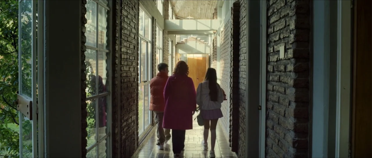 Disconnected 2 (2025) movie still: fullbody — A long interior hallway features exposed brick walls and a floor-to-ceiling glass window wall. Three…