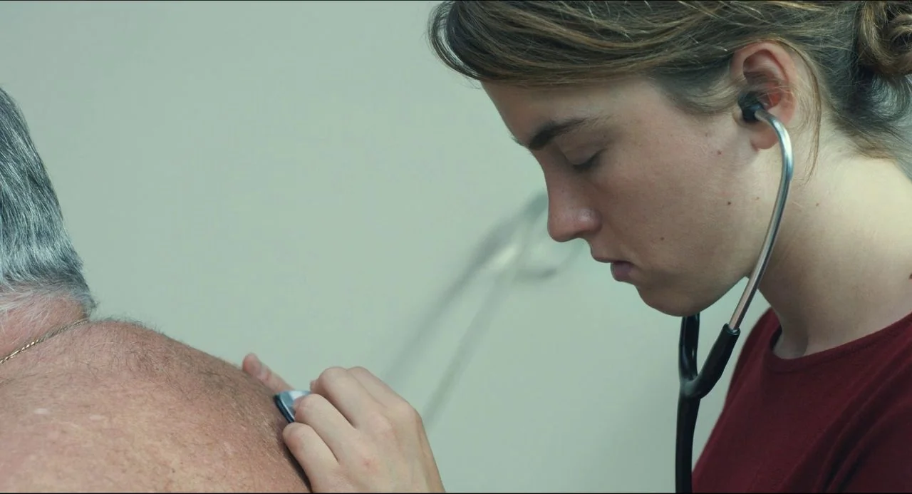 The Unknown Girl (2016) movie still: medium — A clinical examination room has plain, light-colored walls behind the subjects. A young woman uses a…