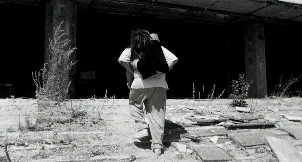 Container (2006) movie still: fullbody — A figure carries someone on their back toward the dark entrance of a derelict building. The ground is…