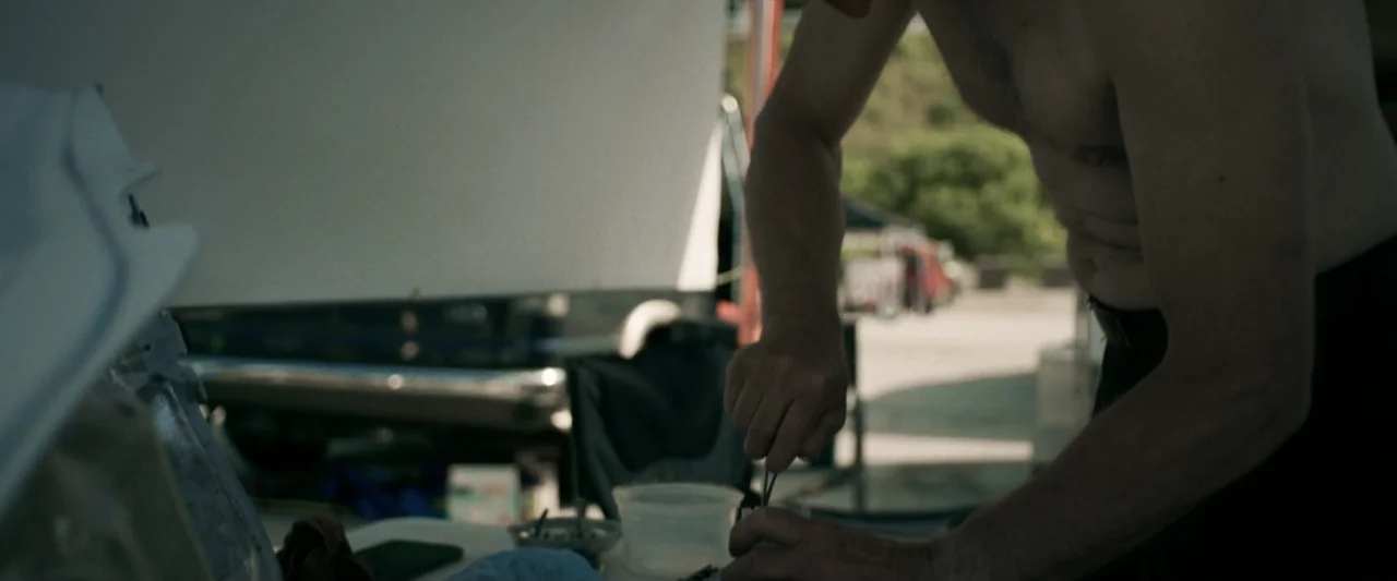 One Fast Move (2024) movie still: medium — An outdoor area features a table cluttered with various tools and containers. A shirtless man leans…