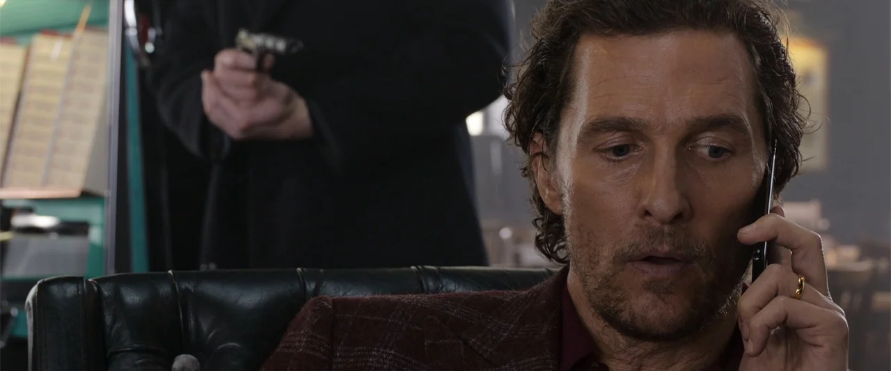 The Gentlemen (2020) movie still: closeup — A man sits in a leather booth at a diner talking on his phone. A second person stands behind him in the…