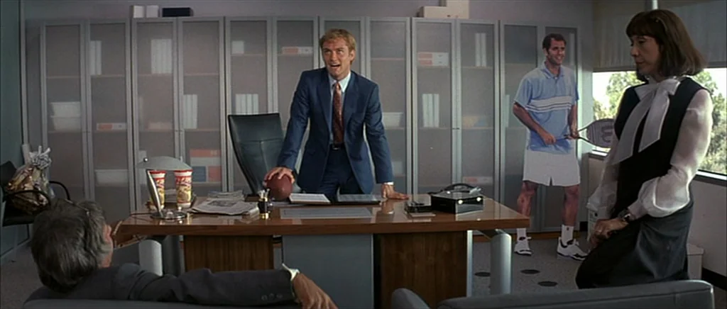 I Heart Huckabees (2004) movie still: wide — A modern corporate workspace features grey filing cabinets, a polished wooden desk, and large windows.…
