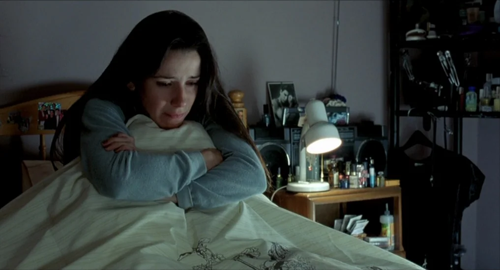 All or Nothing (2002) movie still: medium — A dark, cluttered space with a wooden headboard and a black metal shelving unit. A woman rests in bed…