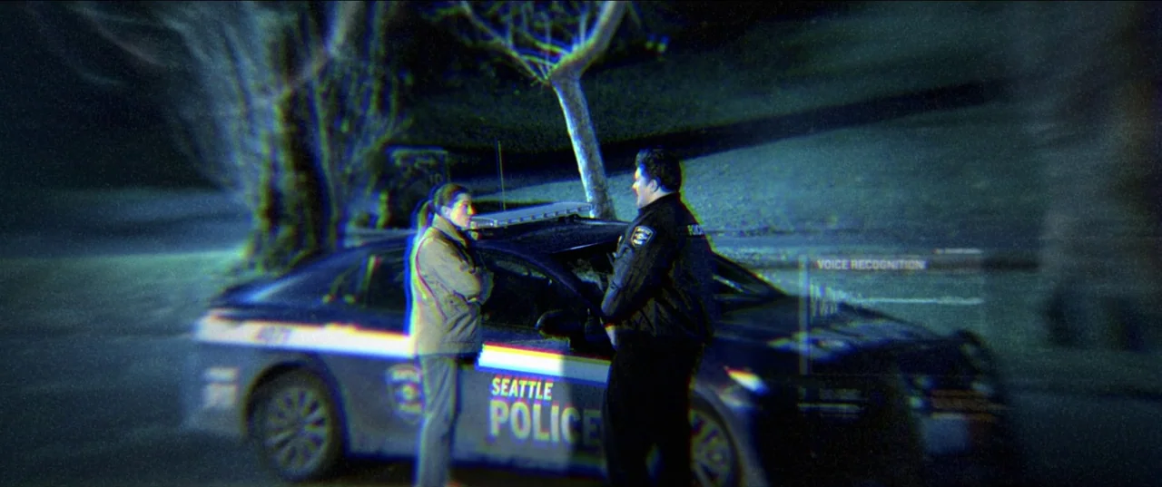 M3gan (2022) movie still: medium — A night scene shows a Seattle police patrol car parked near a large tree. A police officer and a civilian stand…