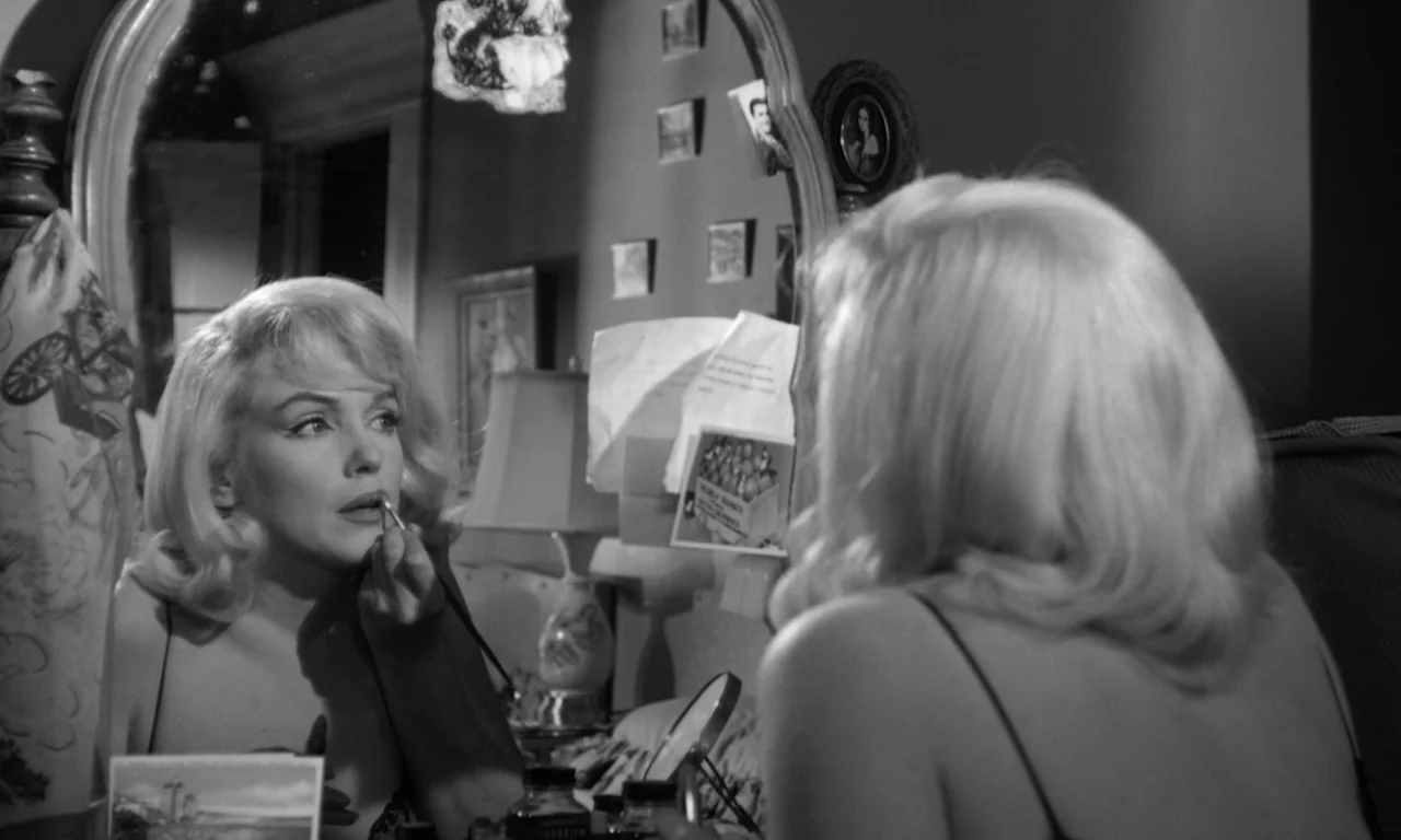 The Misfits (1961) movie still: medium — A woman sits before a vanity mirror in a dimly lit room at night. She holds a small brush and applies…