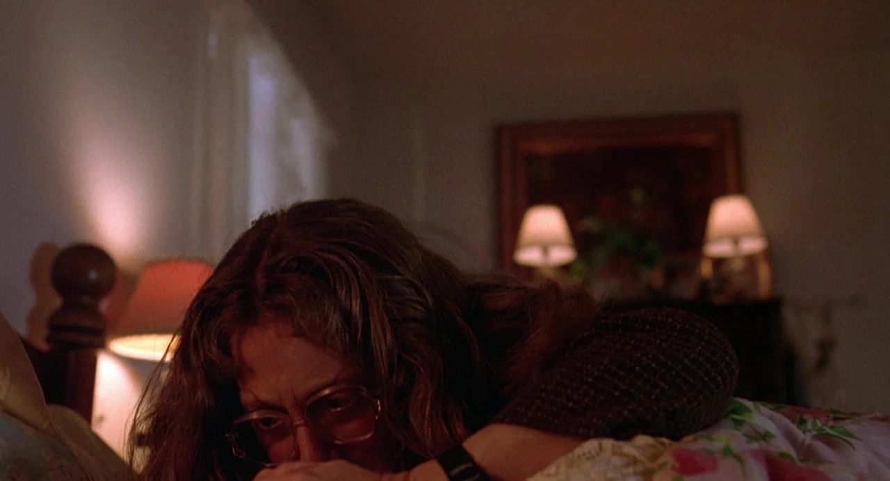 Office Killer (1997) movie still: closeup — A dimly lit room features a wooden bed frame and bedside lamps casting glows. A woman with long…