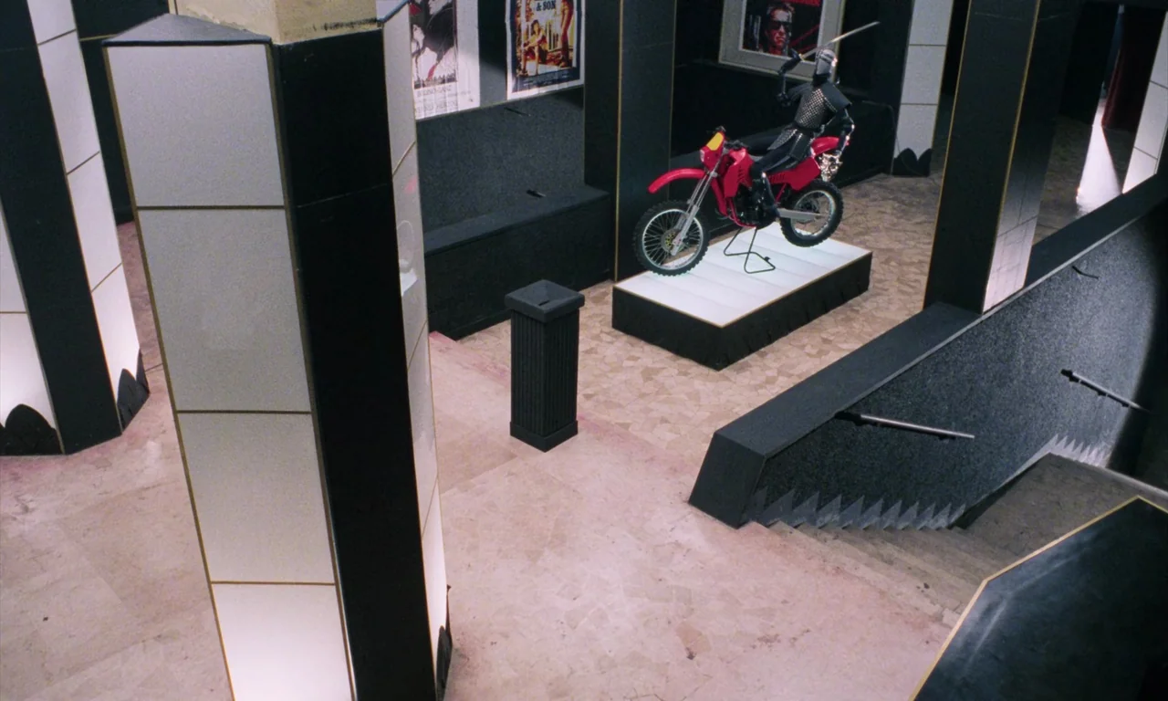 Demons (1985) movie still: wide — A spacious museum or exhibit hall features black and white geometric pillars and walls. A bright red motorcycle…