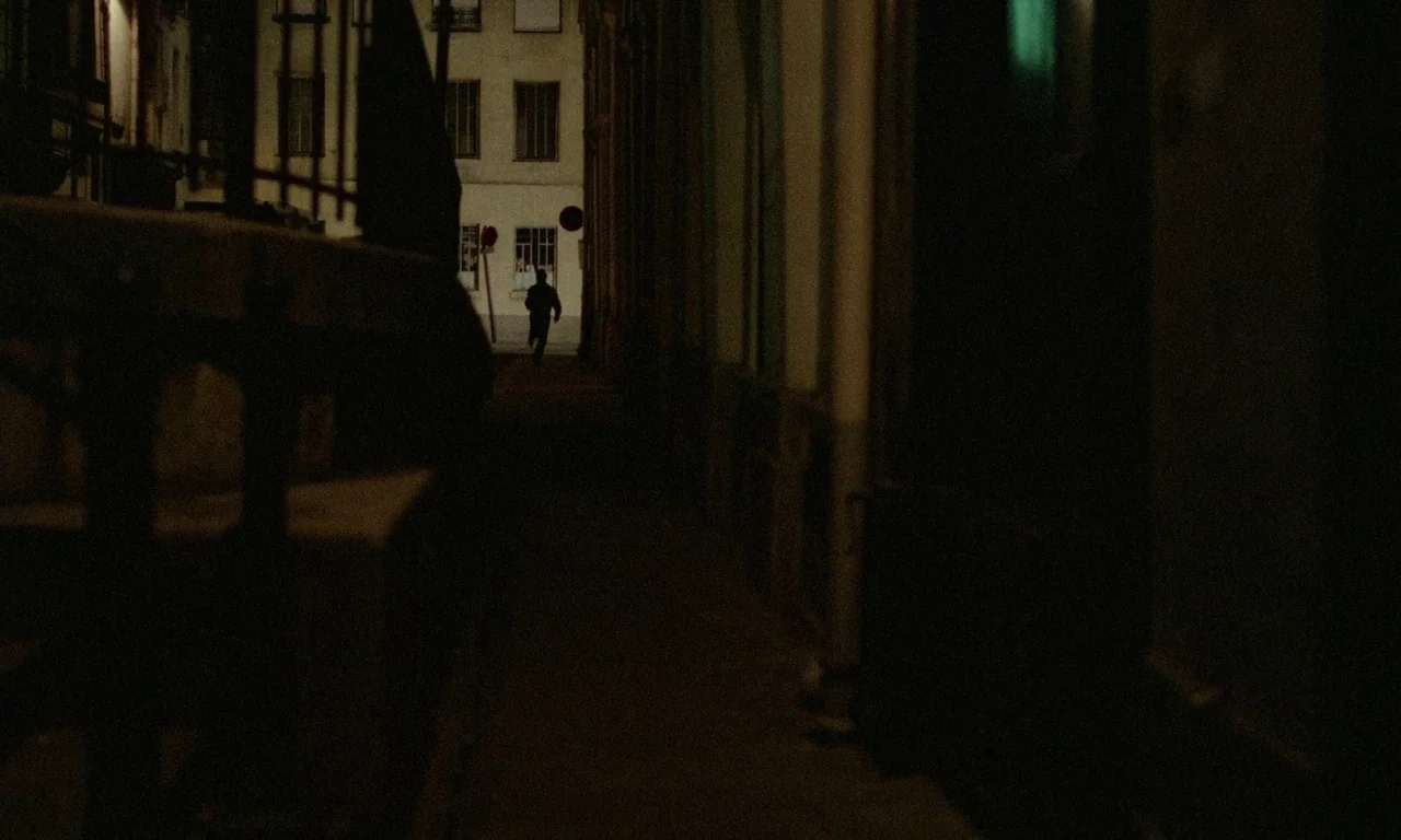 The Margin (1976) movie still: wide — A dark city alleyway leads to a brighter building with visible windows at night. A lone figure in silhouette…