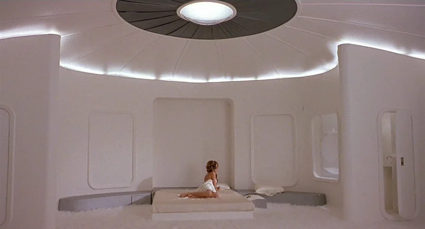 CQ (2001) movie still: wide — A circular futuristic room features white curved walls and a central ceiling light fixture. A woman in a white wrap…
