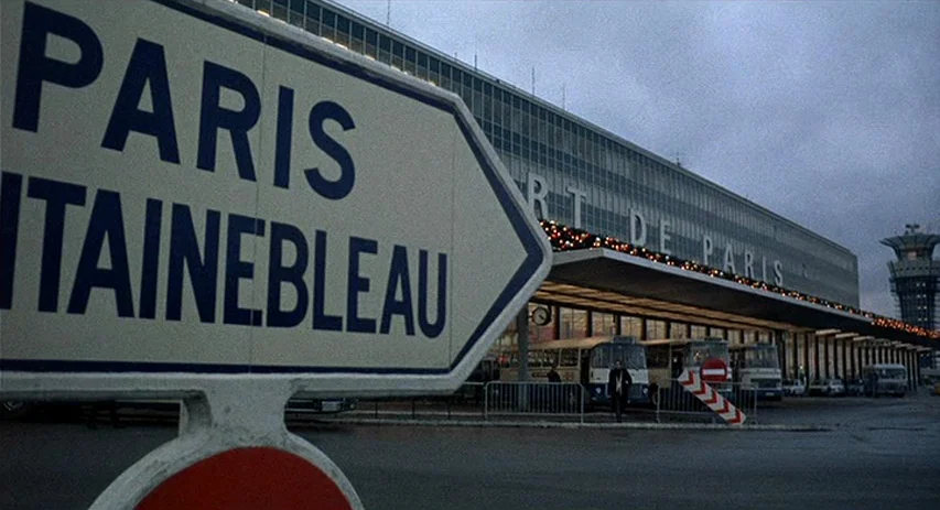 Perversion Story (1969) movie still: wide — A modernist airport exterior features a large glass-walled terminal under a cloudy sky. A prominent…