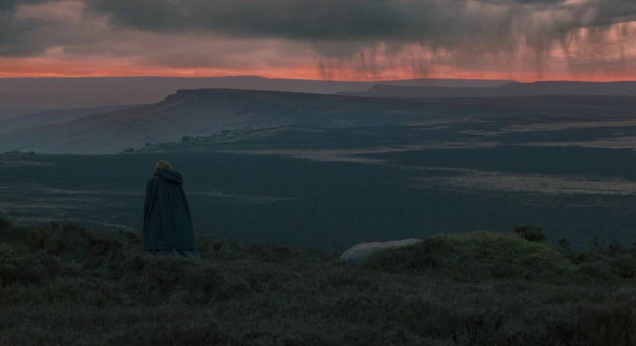 Jane Eyre (2011) movie still: wide — A vast, dark moorland stretches toward a distant ridge under a rain-streaked sunset sky. A lone person in a…