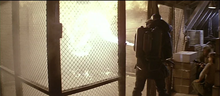 The Thing (1982) movie still: medium — A man with a flamethrower stands inside an industrial storage cage area. He projects a stream of fire…