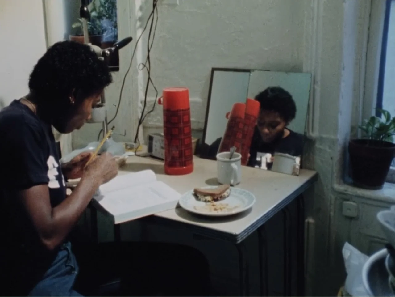 Born in Flames (1983) movie still: medium — A cluttered tabletop serves as a workspace in a sparsely furnished room. A woman sits writing in a…