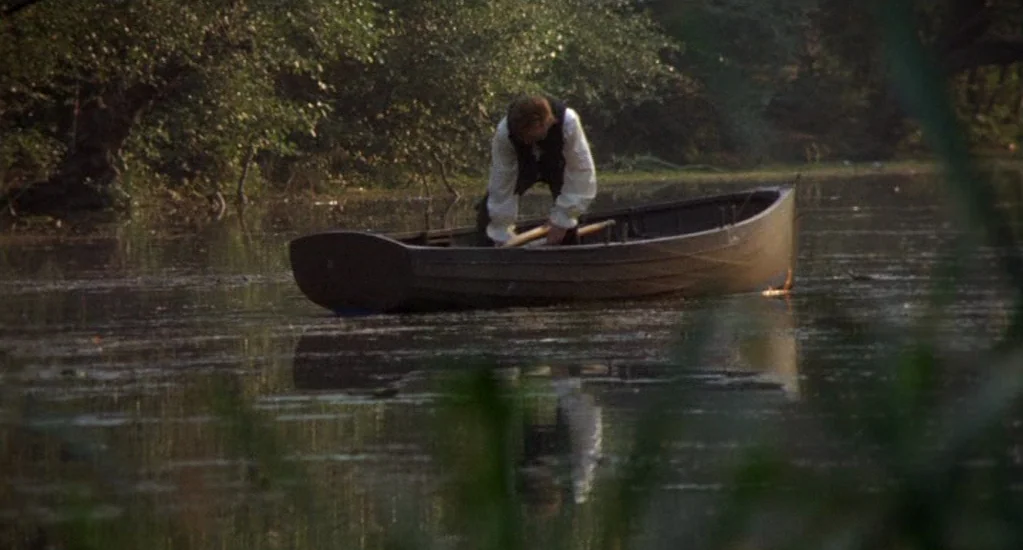 Demons of the Mind (1972) movie still: wide — A calm, stagnant water body is surrounded by dense, overgrown vegetation and dark trees. A man wearing…
