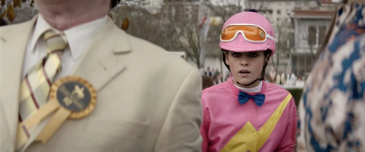 Charlie's Angels (2019) movie still: medium — An outdoor horse racing event features a crowd gathered in a paved area. A female jockey wearing pink…