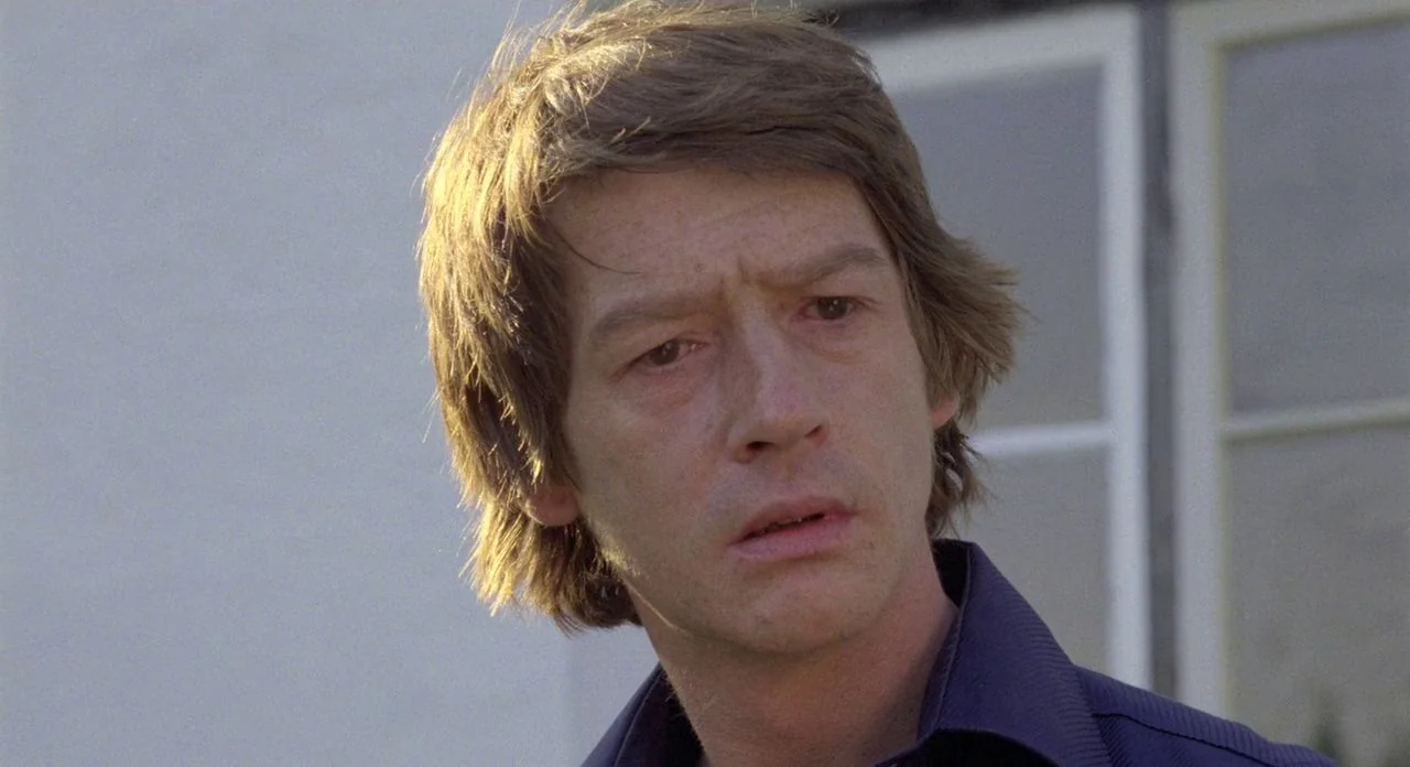 The Shout (1978) movie still: closeup — A man with light brown hair stands outdoors in front of a white wall. The subject looks off-camera while…