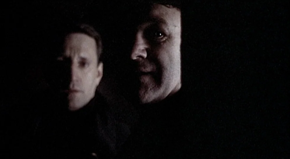 The French Connection (1971) movie still: medium — Two men stand in a dimly lit, dark, and featureless space. They stare ahead with their faces…