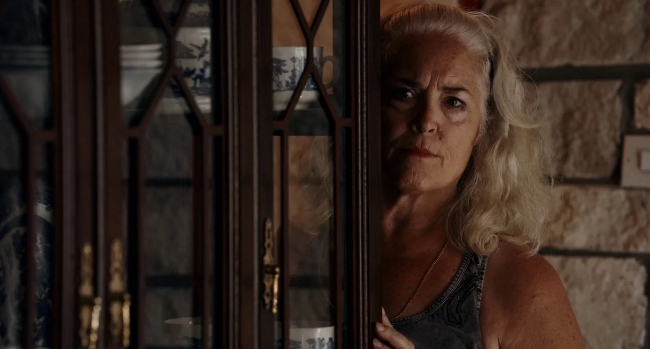 Krisha (2015) movie still: closeup — A woman stands partly obscured behind the wooden frame of a china cabinet. She peers through the glass panes at…