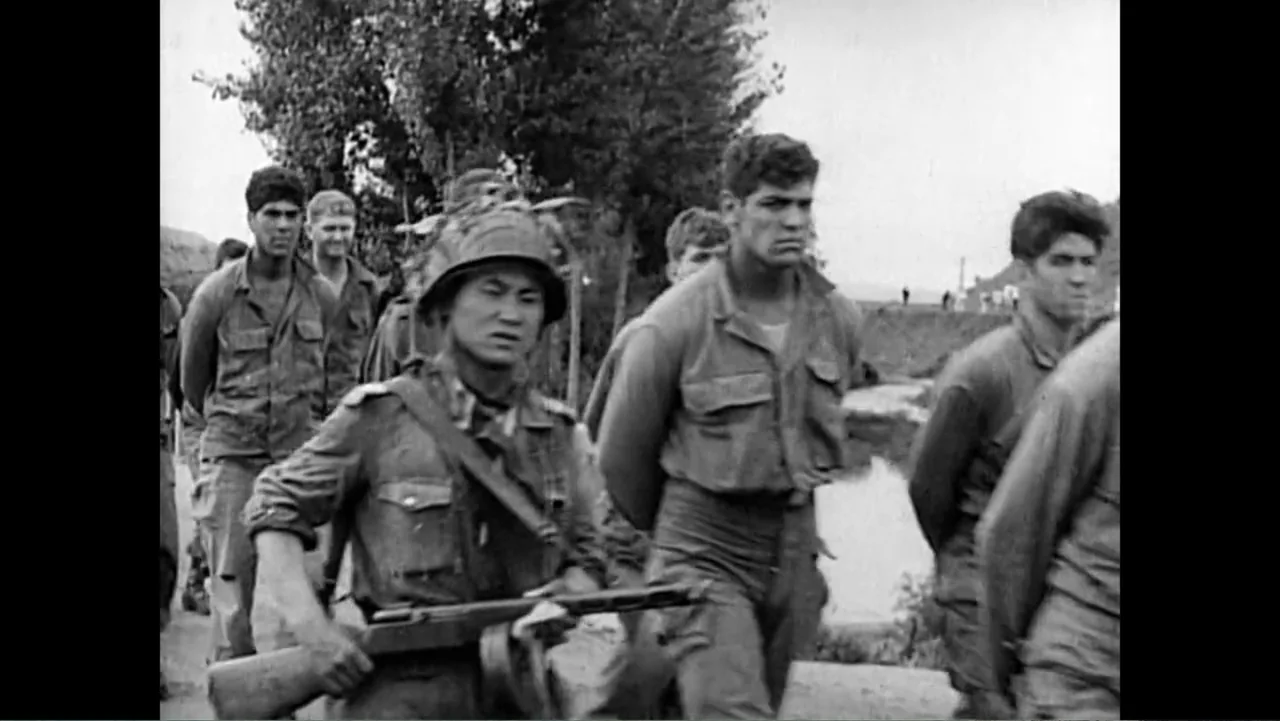 The Forever Prisoner (2021) movie still: medium — A group of soldiers walks along a dirt path flanked by trees and foliage. One soldier in the…