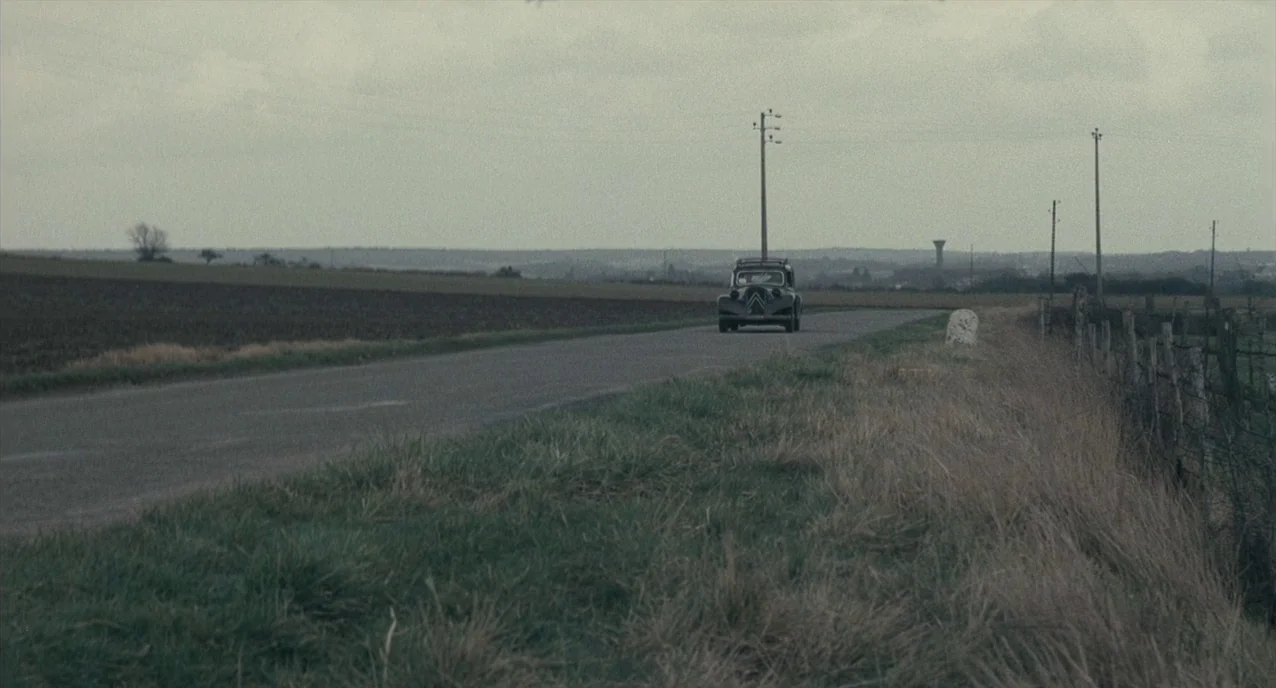 The Army of Shadows (1969) movie still: wide — A rural landscape features a paved road flanked by brown fields and tall grass. An old black sedan…