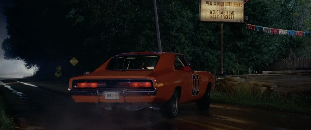 The Dukes of Hazzard (2005) movie still: wide — A rural asphalt road at night with an orange muscle car parked prominently. A tall sign advertising…