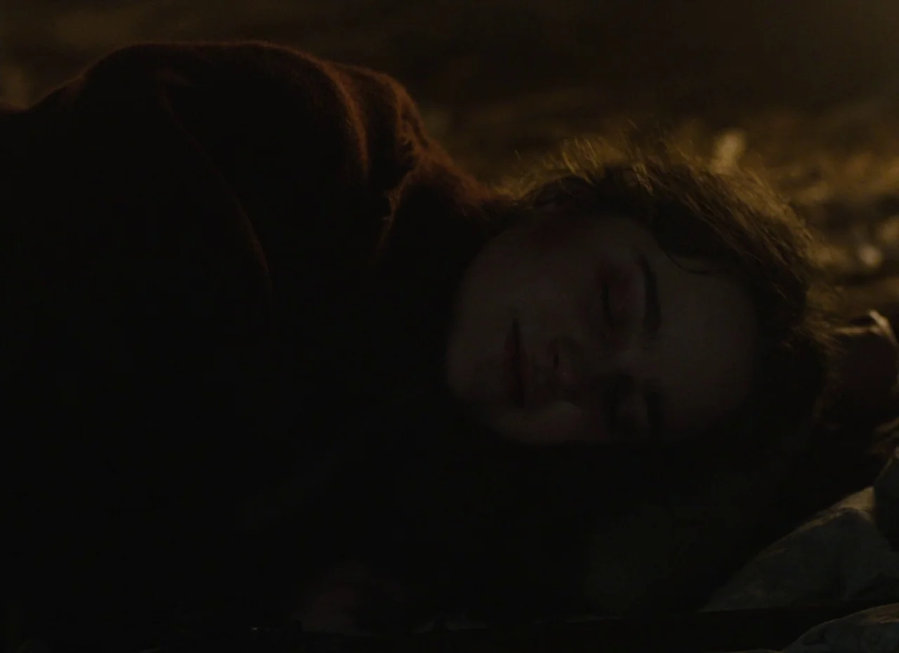 The Nightingale (2018) movie still: closeup — A dimly lit room provides a dark, shadowed environment. A person lies with their head resting against…