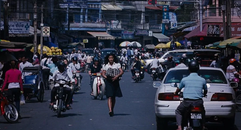 Almayer’s Folly (2011) movie still: wide — A dense urban intersection in Cambodia is filled with cars, motorbikes, and pedestrians. A woman crosses…