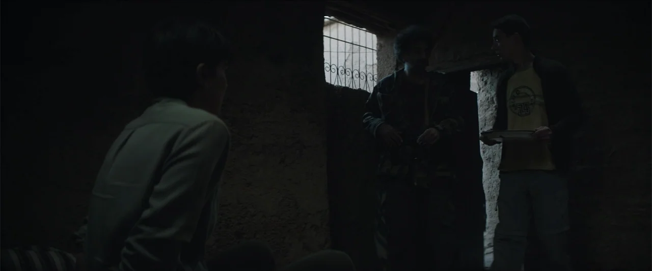 Redemption Day (2021) movie still: medium — A dark, unfinished interior space has rough textured walls and a small barred window. Two young men…