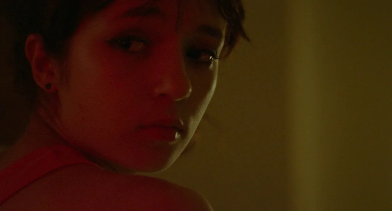 It Felt Like Love (2013) movie still: closeup — A dimly lit interior room at night with warm red lighting. A young woman turns her head to look…