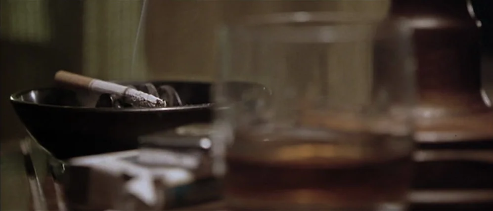Apt Pupil (1998) movie still: closeup — A close-up of a dark ashtray holding a single cigarette next to a glass. An amber-colored drink fills the…