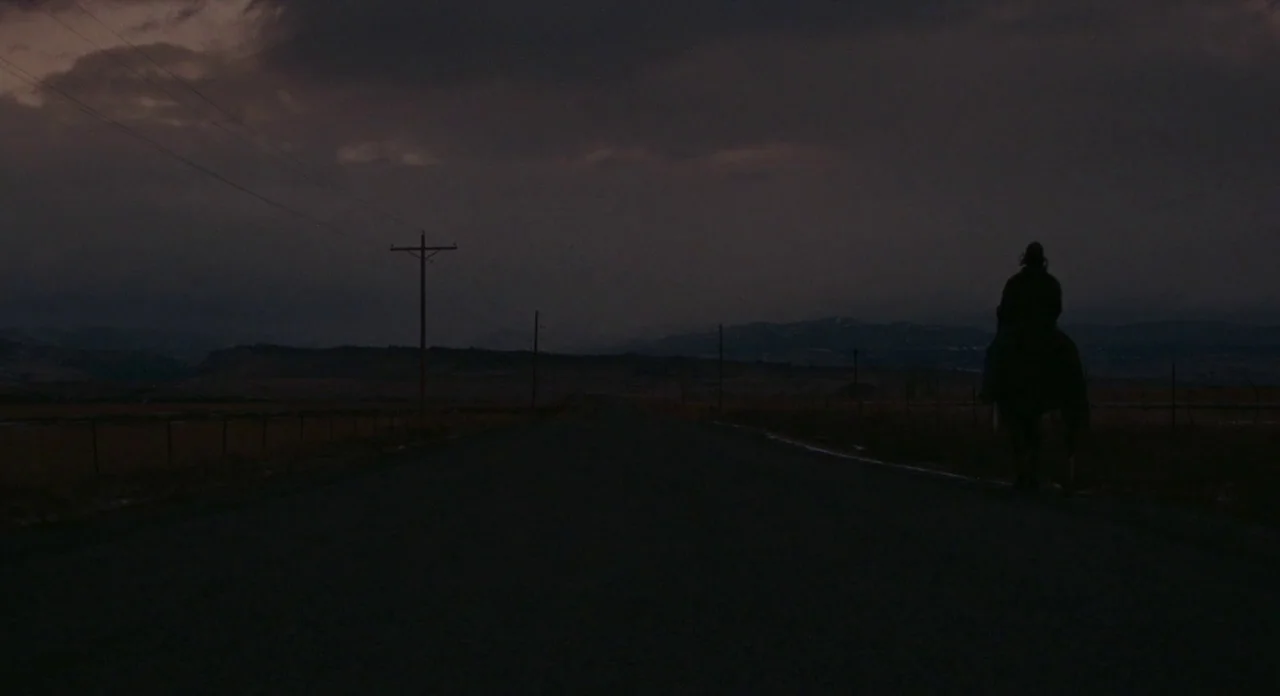 Certain Women (2016) movie still: wide — A dark, empty asphalt road stretches toward distant hills under a cloudy evening sky. A rider on horseback…
