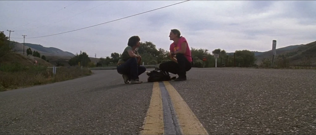 Death Proof movie still: wide — A rural paved road stretches through a valley under a cloudy sky. Two women crouch facing each other on the yellow…