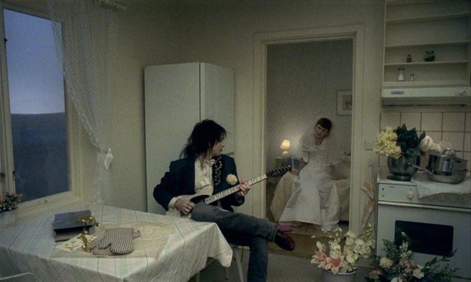 You, The Living (2007) movie still: medium — A sparely furnished apartment shows a dining area in the foreground and bedroom behind. A person plays…