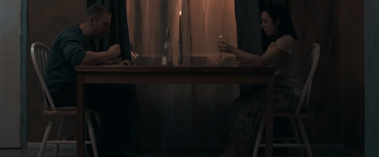 Anomaly (2006) movie still: medium — A dimly lit room with a wooden table placed in front of a window. A man and a woman sit facing each other while…