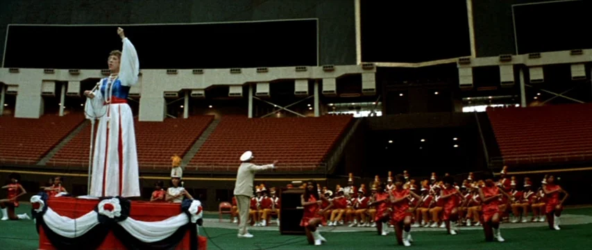 Brewster McCloud (1970) movie still: wide — A vast empty sports stadium features rows of red plastic seats and a green field. A woman stands on a…