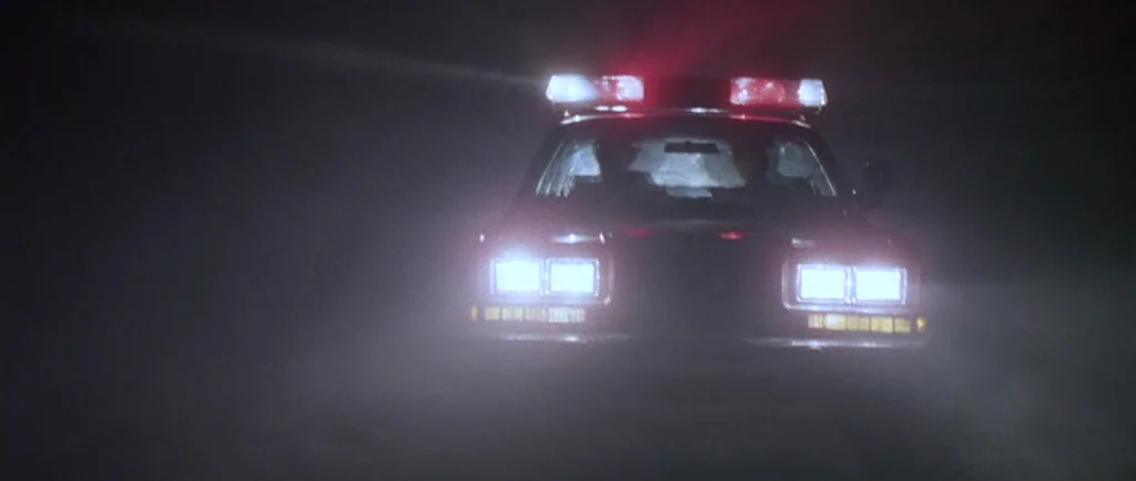 Manhunter (1986) movie still: wide — A dark, empty road is occupied only by the front of a police car. The vehicle is stationary with its bright…