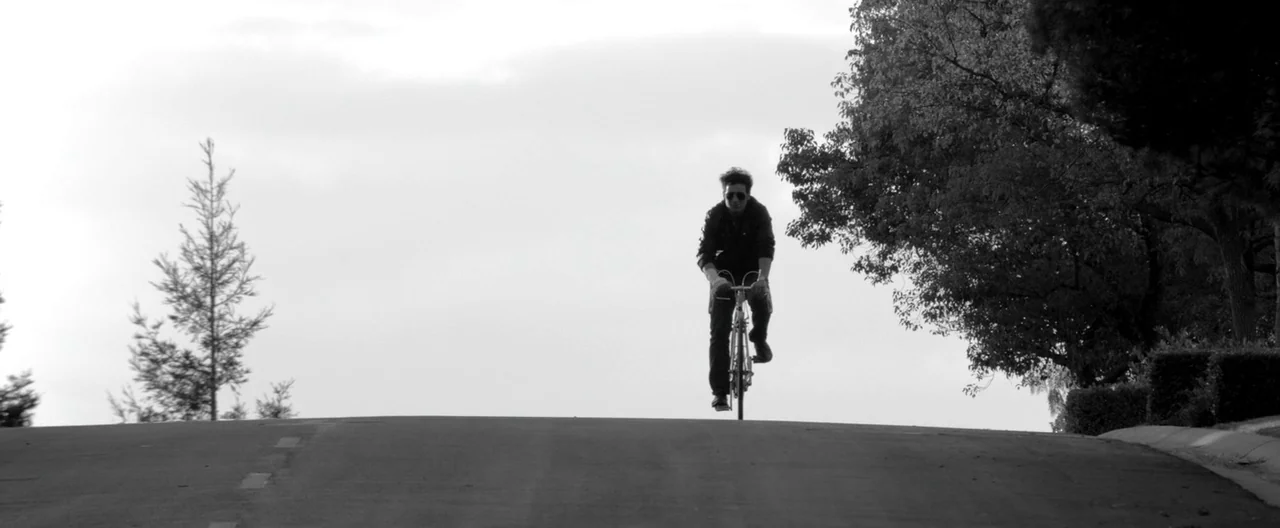 A Girl Walks Home Alone at Night (2014) movie still: wide — An asphalt road rises to a horizon line against a bright overcast sky. A man rides a…