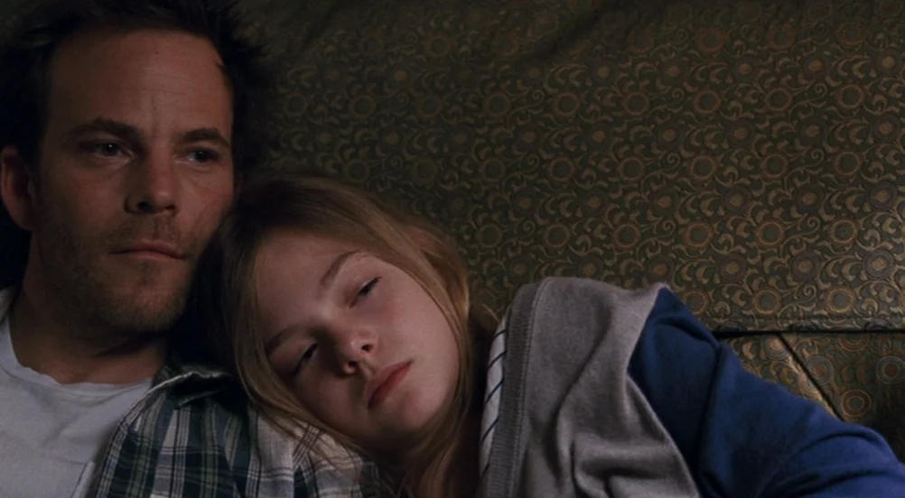 Somewhere (2010) movie still: medium — A dimly lit interior space features a man and a child on a couch. The man rests his head near the girl while…