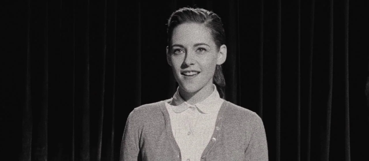 Seberg (2019) movie still: medium — A dark, plain curtain forms the backdrop behind a woman in a cardigan. The woman stands facing forward in a…