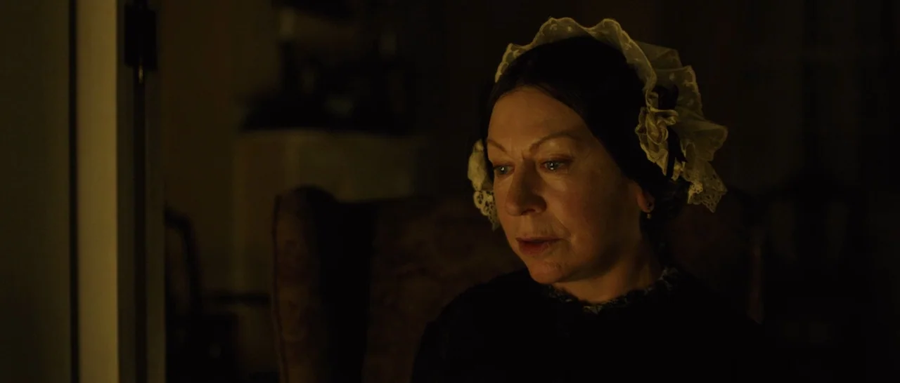 A Quiet Passion (2016) movie still: closeup — A dimly lit room featuring a woman in 19th-century attire. She sits in a dark chair while looking…
