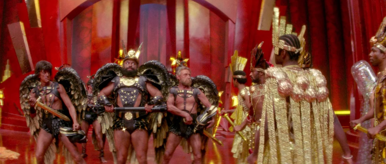 Flash Gordon (1980) movie still: wide — A high-ceilinged room with deep red walls and decorative gold geometric panels. Several winged warriors in…