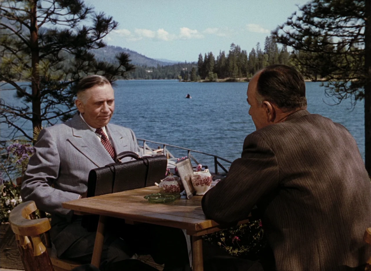 Leave Her To Heaven (1945) movie still: medium — A wooden table is set on a patio overlooking a calm lake surrounded by pine trees. Two middle-aged…