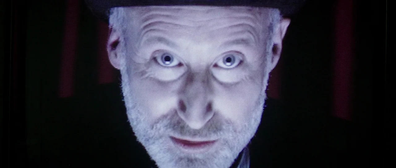 The Babadook (2014) movie still: closeup — A man's face fills the frame, appearing on a grainy low-resolution television monitor. Dark red vertical…