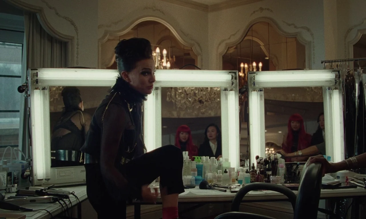 Vox Lux (2018) movie still: medium — A well-lit dressing room features large vanity mirrors with surrounding fluorescent bulb strips. A woman sits…