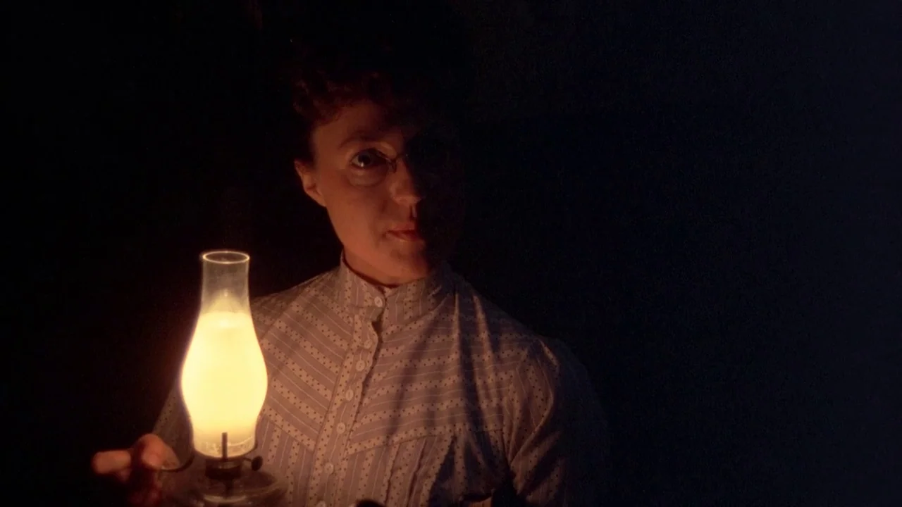 Picnic at Hanging Rock (1975) movie still: medium — A woman in a pinstriped blouse stands in an otherwise dark room. She holds a small, glowing oil…