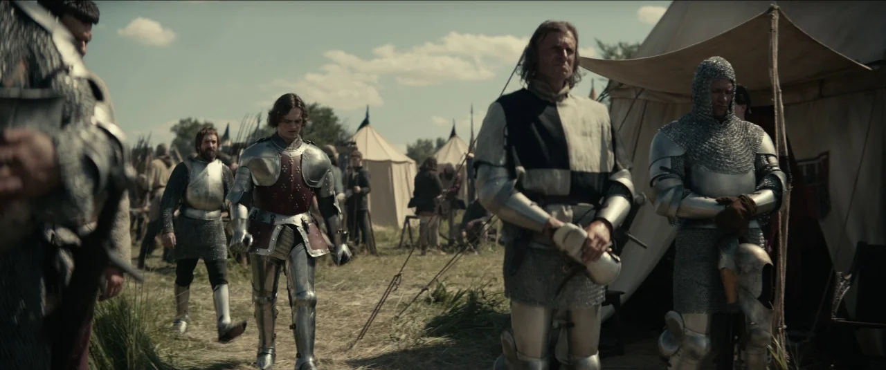 The King movie still: fullbody — A field encampment features several canvas tents arranged on dry grass under blue skies. Armored knights in…