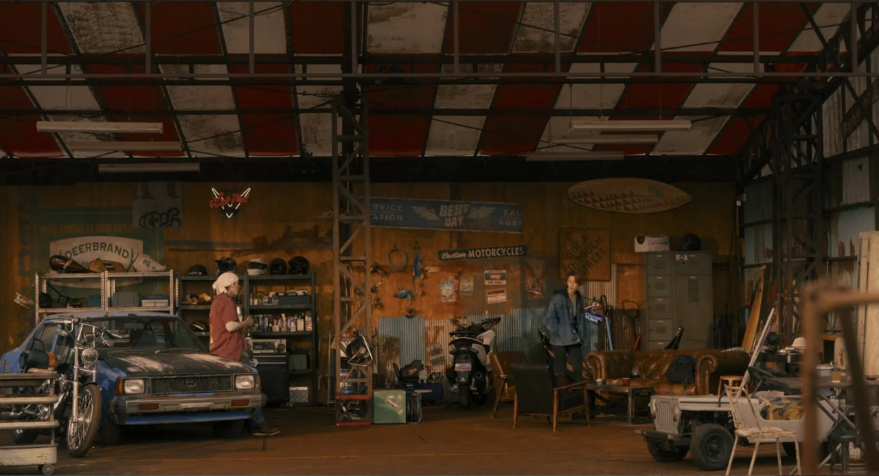 Knuckle Girl (2023) movie still: wide — A spacious, cluttered industrial garage features multiple motorcycles, a car, and various repair tools. Two…
