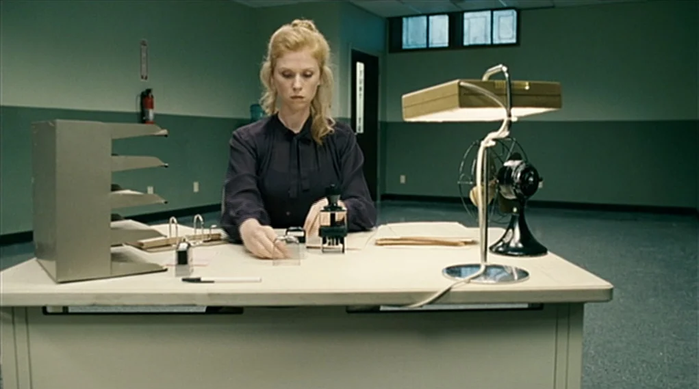 Visioneers (2008) movie still: medium — A utilitarian office space with mint green walls and a gray linoleum floor. A woman in a dark blouse sits at…